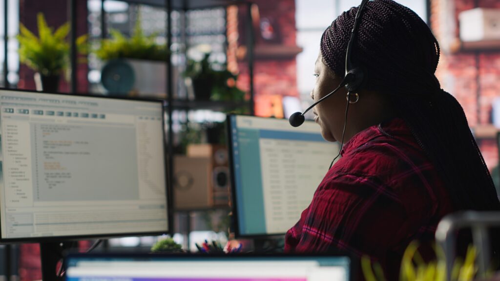 A woman in a headset works at a desk, using two computers simultaneously.