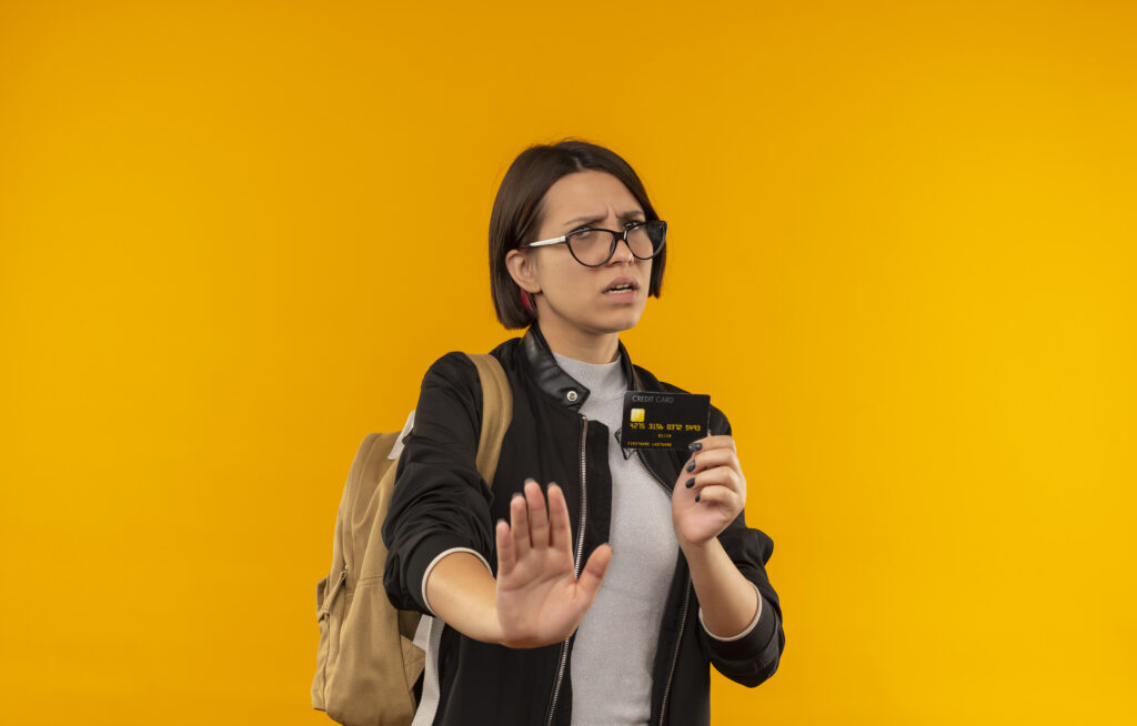 unpleased young student girl wearing glasses and back bag holding credit card gesturing stop isolated on orange background with copy space