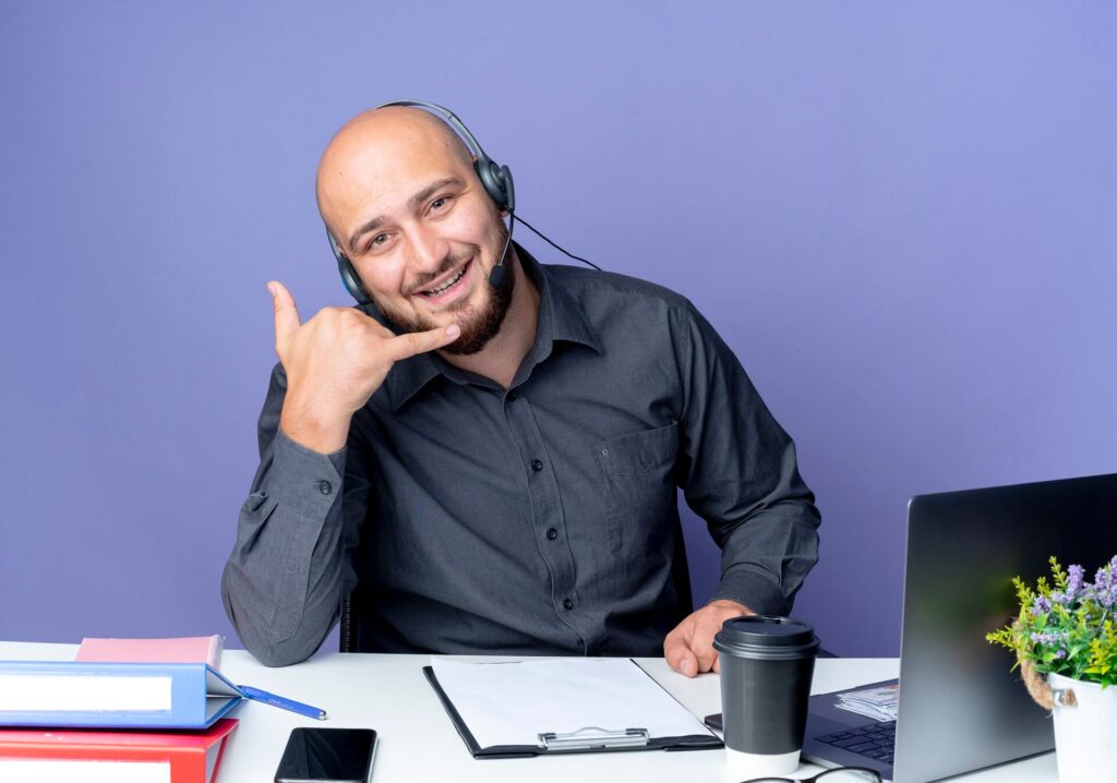 joyful-young-bald-call-center-man-wearing-headset-sitting-desk-with-work-tools-doing-call-gesture-isolated-purple